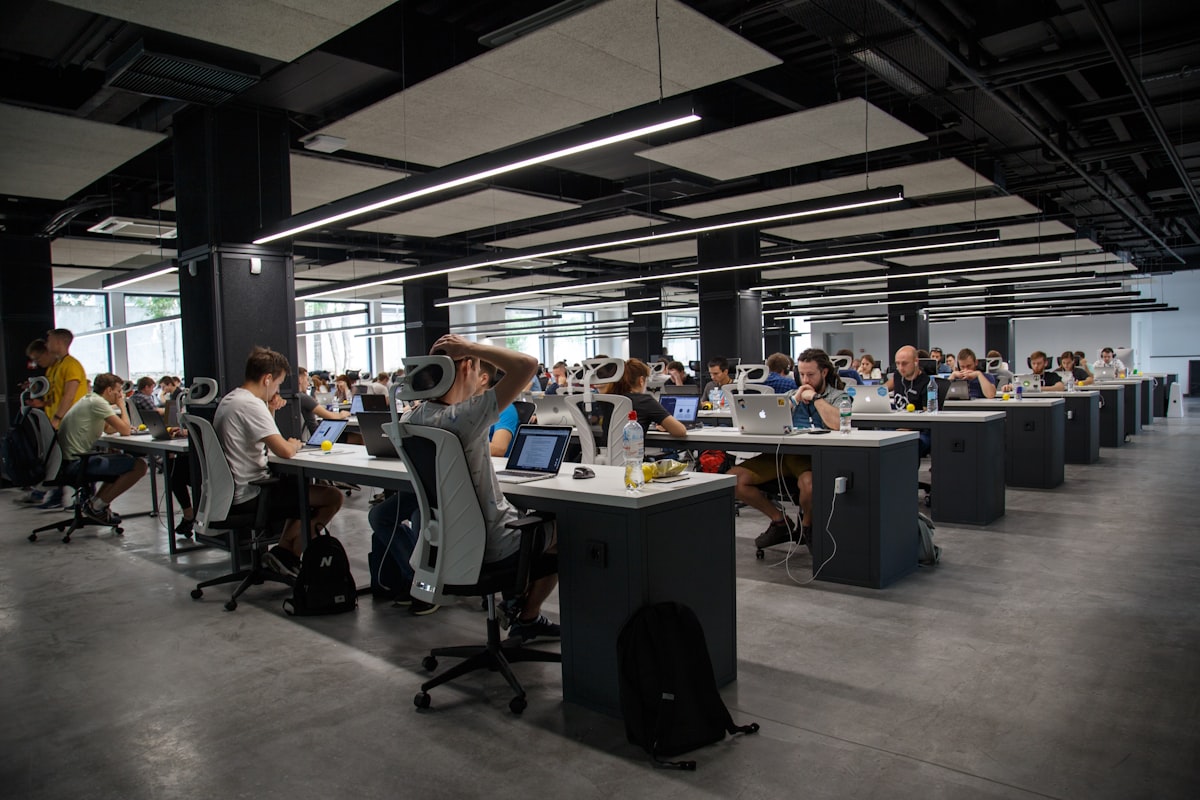 Grand open space professionnel avec plusieurs rangées de collaborateurs travaillant sur des ordinateurs portables sous un plafond industriel éclairé de néons linéaires