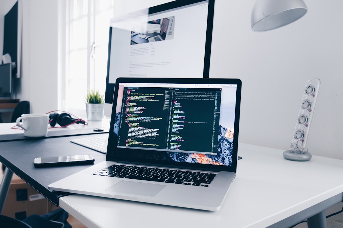 Ordinateur portable posé sur un bureau blanc affichant du code, avec un second écran en arrière-plan dans un bureau clair