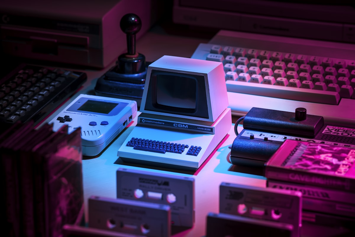 Collection d'ordinateurs et de consoles vintage — un Commodore CBM, une Game Boy et des cassettes — disposés sur une surface éclairée par une lumière neon rose et violette